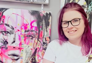 girl with pink hair and white shirt smiling in front of female portrait on canvas